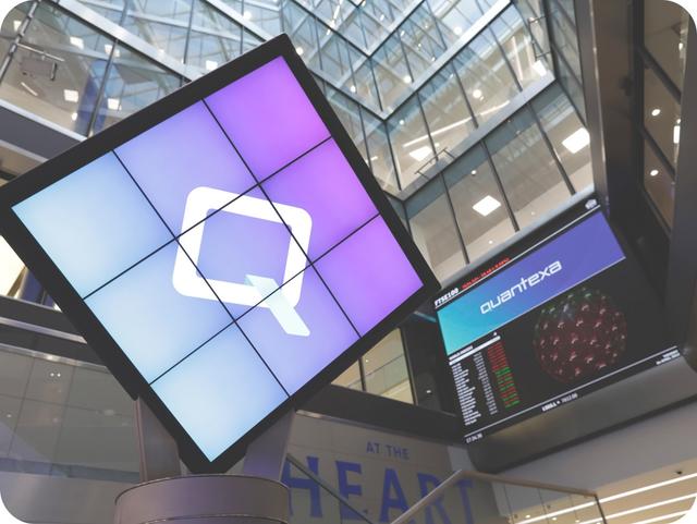 Digital display with a logo on a multi-screen setup in a modern building interior, alongside a stock market ticker showing "quantexa."