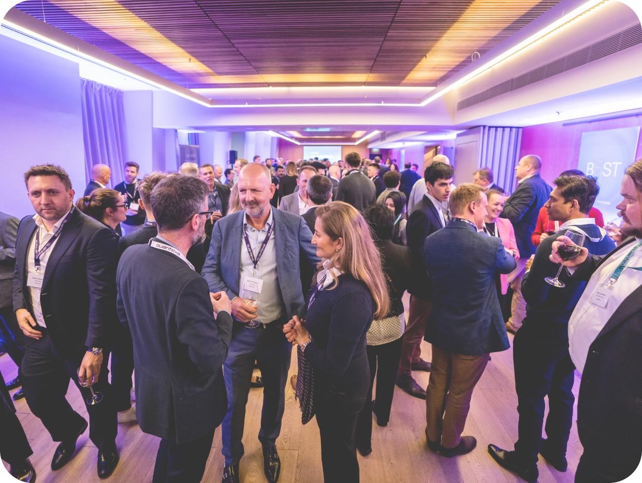 A large group of people in formal attire engage in conversations at a networking event in a well-lit room with purple ambient lighting.