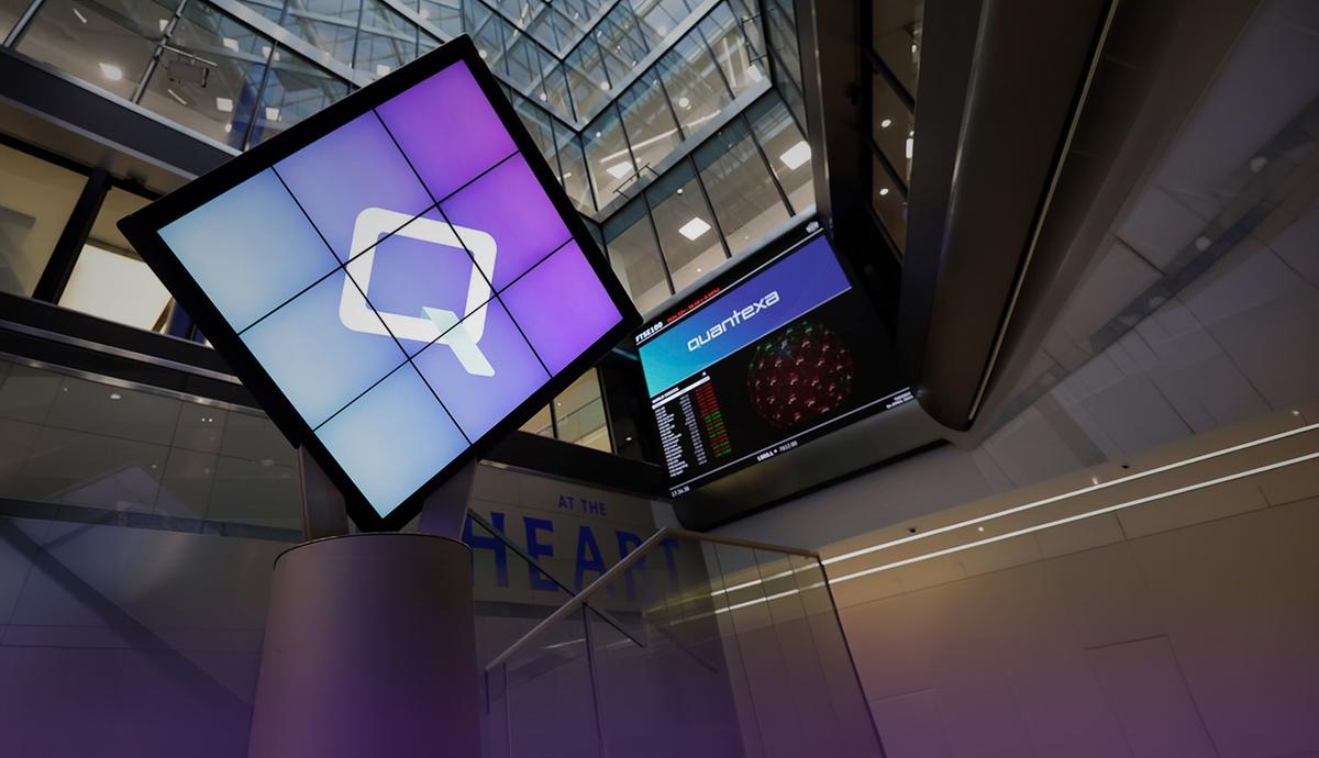 A modern office atrium with a digital display featuring a geometric logo and a separate screen displaying graphs. Glass and metal architecture.