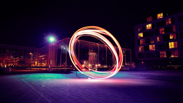 A vibrant light painting circle in a nighttime urban setting, with illuminated buildings in the background.