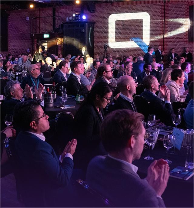 Audience seated at tables, attentively watching a presentation in a dimly lit venue with exposed brick walls and stage lighting.
