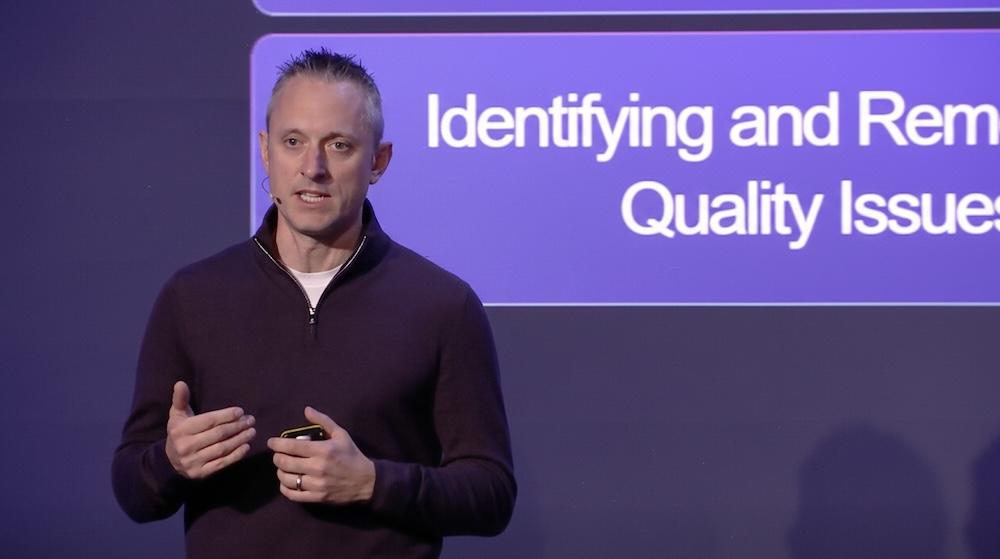 Person speaking on stage about identifying and removing quality issues, standing in front of a large screen with a purple background.