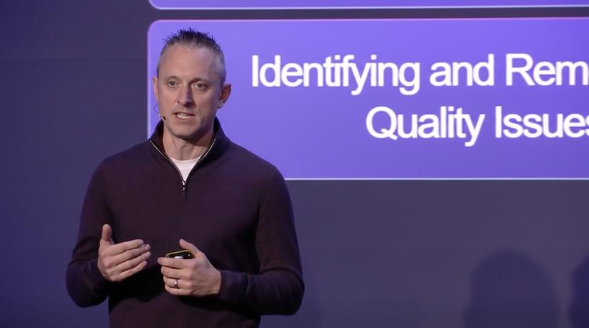 Person speaking on stage about identifying and removing quality issues, standing in front of a large screen with a purple background.