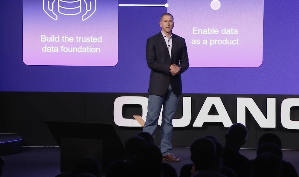 Dan Higgins in a blazer and jeans speaks on stage with a presentation about data foundation and products displayed on the screen behind.