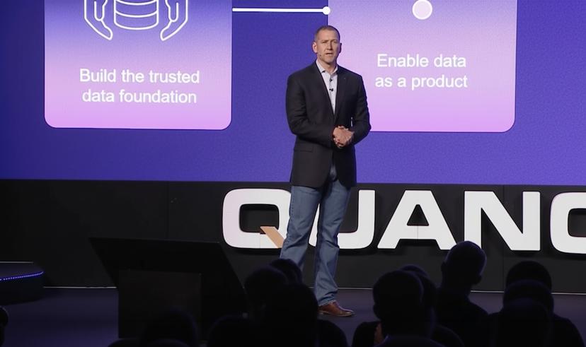 Dan Higgins in a blazer and jeans speaks on stage with a presentation about data foundation and products displayed on the screen behind.