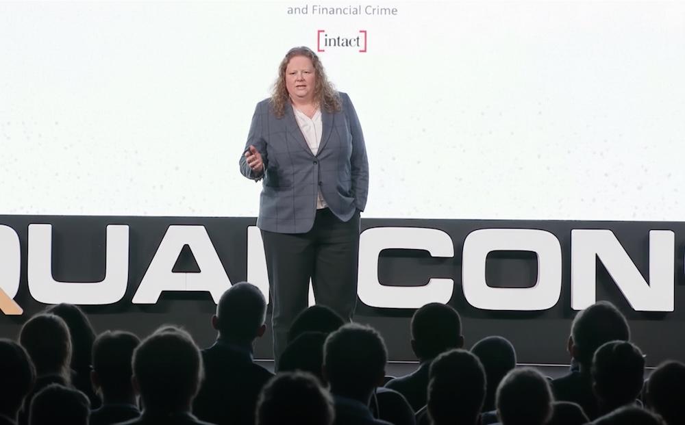 Speaker in a blazer presenting on stage with a large audience, conference graphics in the background.