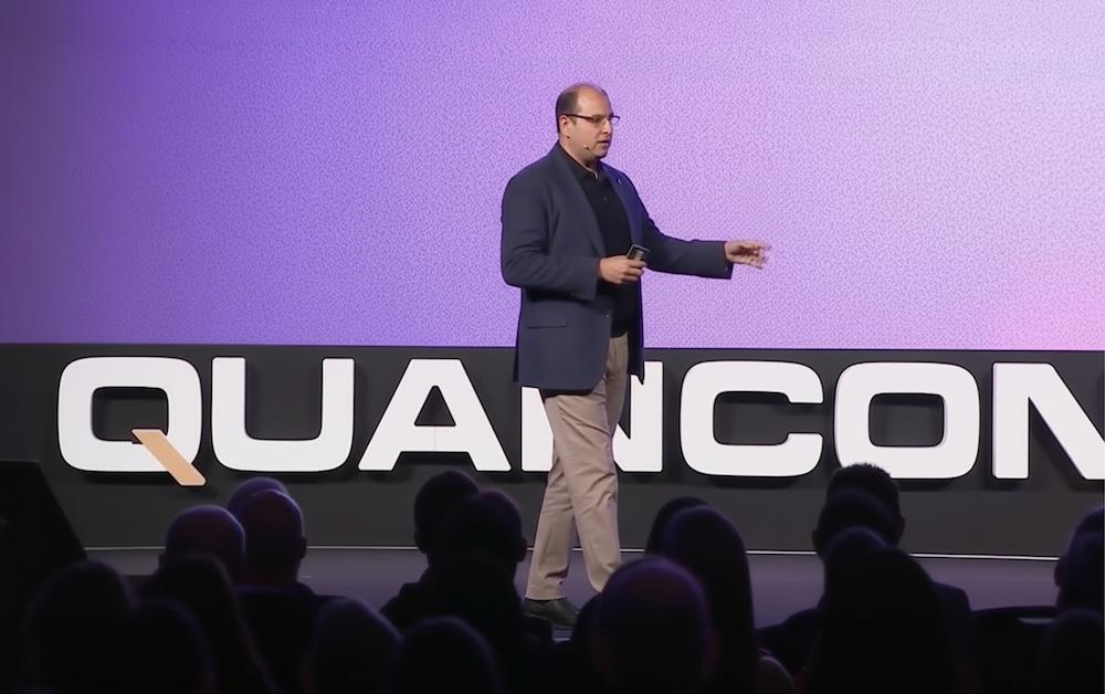 A person in a dark jacket and tan pants speaks on stage, with a purple background and partial "QUANCON" signage behind them.