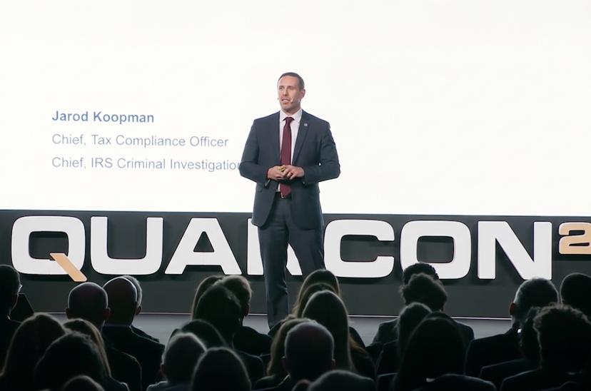 A man in a suit stands on stage speaking at a conference. The background shows the text "QUARCON" and his titles. Audience silhouettes are visible.