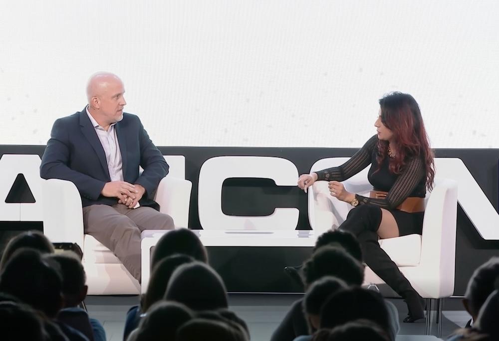 Two people sit on stage engaged in discussion, facing each other. Audience members are visible in the foreground.
