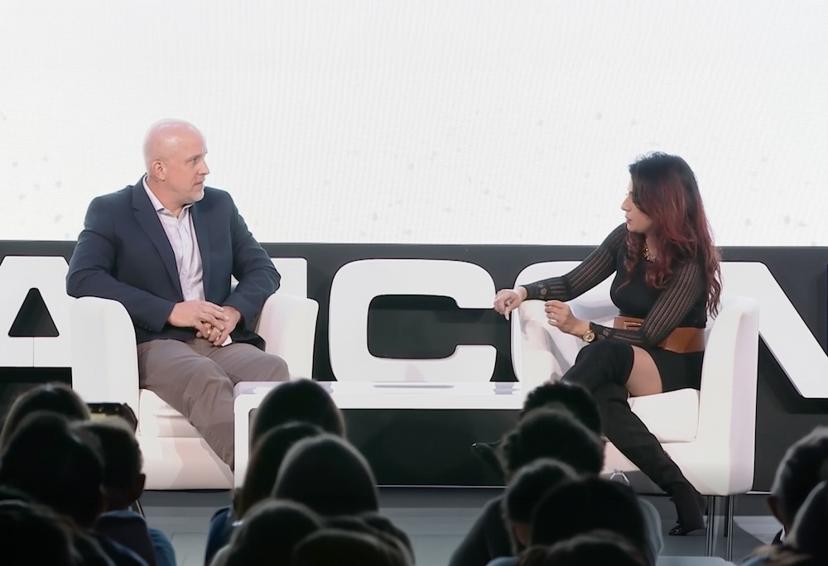 Two people sit on stage engaged in discussion, facing each other. Audience members are visible in the foreground.