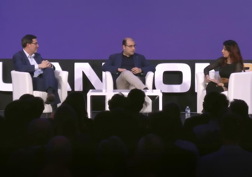 Three people sit on stage discussing in armchairs against a blue backdrop, with an audience in the foreground.