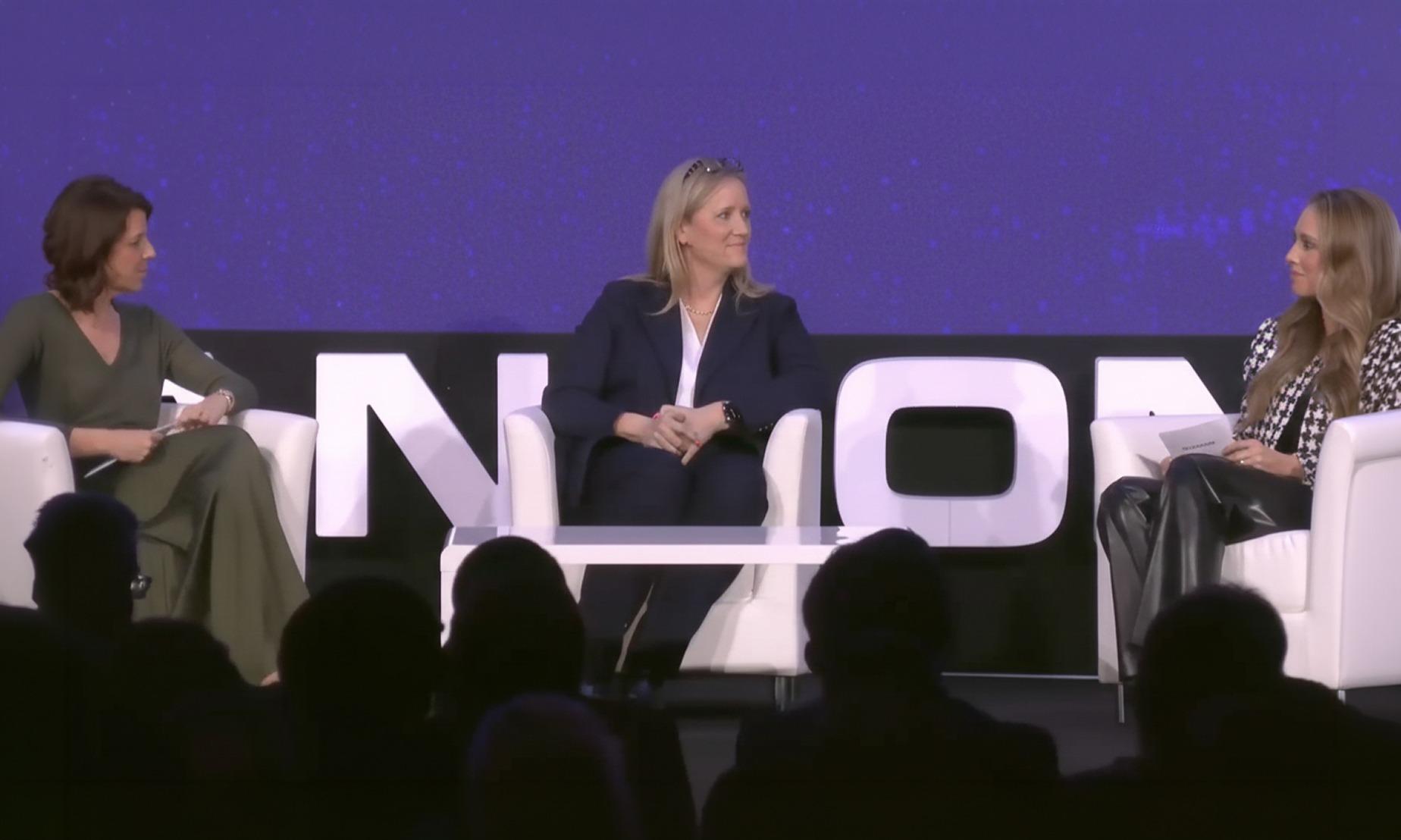Three women sit on stage in chairs, engaged in a panel discussion against a backdrop with large text. One holds notes, and another is speaking.