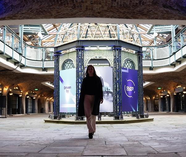 Person walking in a spacious, industrial-style hall with large digital screens and exposed brick arches.