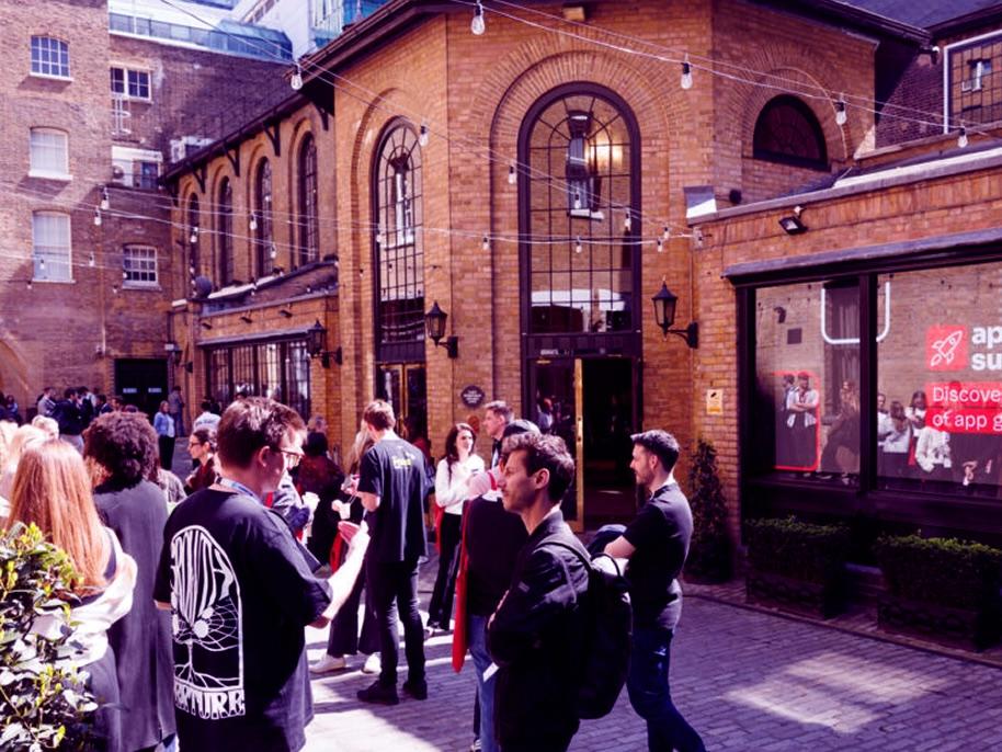 People socializing in a courtyard with brick buildings, string lights, and a poster on the window. Some are holding drinks and chatting.